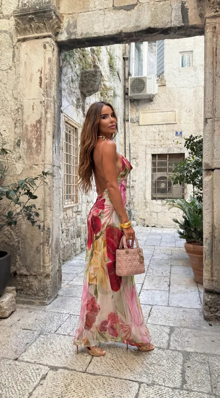 Side view of the floral maxi dress showing its open back, thin beaded straps and fitted silhouette in a stone archway