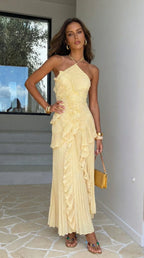 Model in pale yellow pleated halter maxi dress with ruffle detail, hand on hip, standing by a window