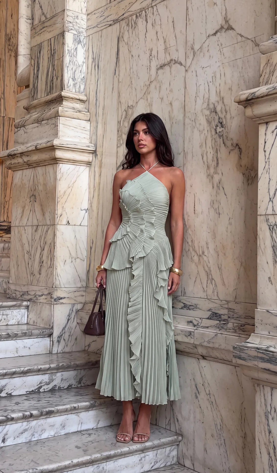 Model standing against architectural backdrop wearing the yellow pleated halter dress with gold jewellery