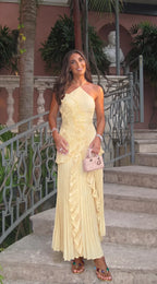 Woman smiling on stone steps wearing yellow pleated halter dress with cascading ruffle and pink handbag