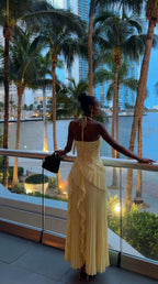 Back view of model on balcony overlooking palm trees, wearing pale yellow pleated halter maxi dress