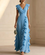 Woman in a dusty blue ruffle maxi dress with V-neckline and flutter sleeves, carrying a small handbag