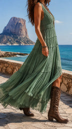Side view of the flowing sage green tiered maxi dress with brown cowboy boots, walking along a stone coastal path