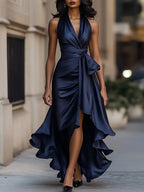 Model wears a navy blue satin halterneck wrap maxi dress with tie waist and ruffled high-low hem on a city street