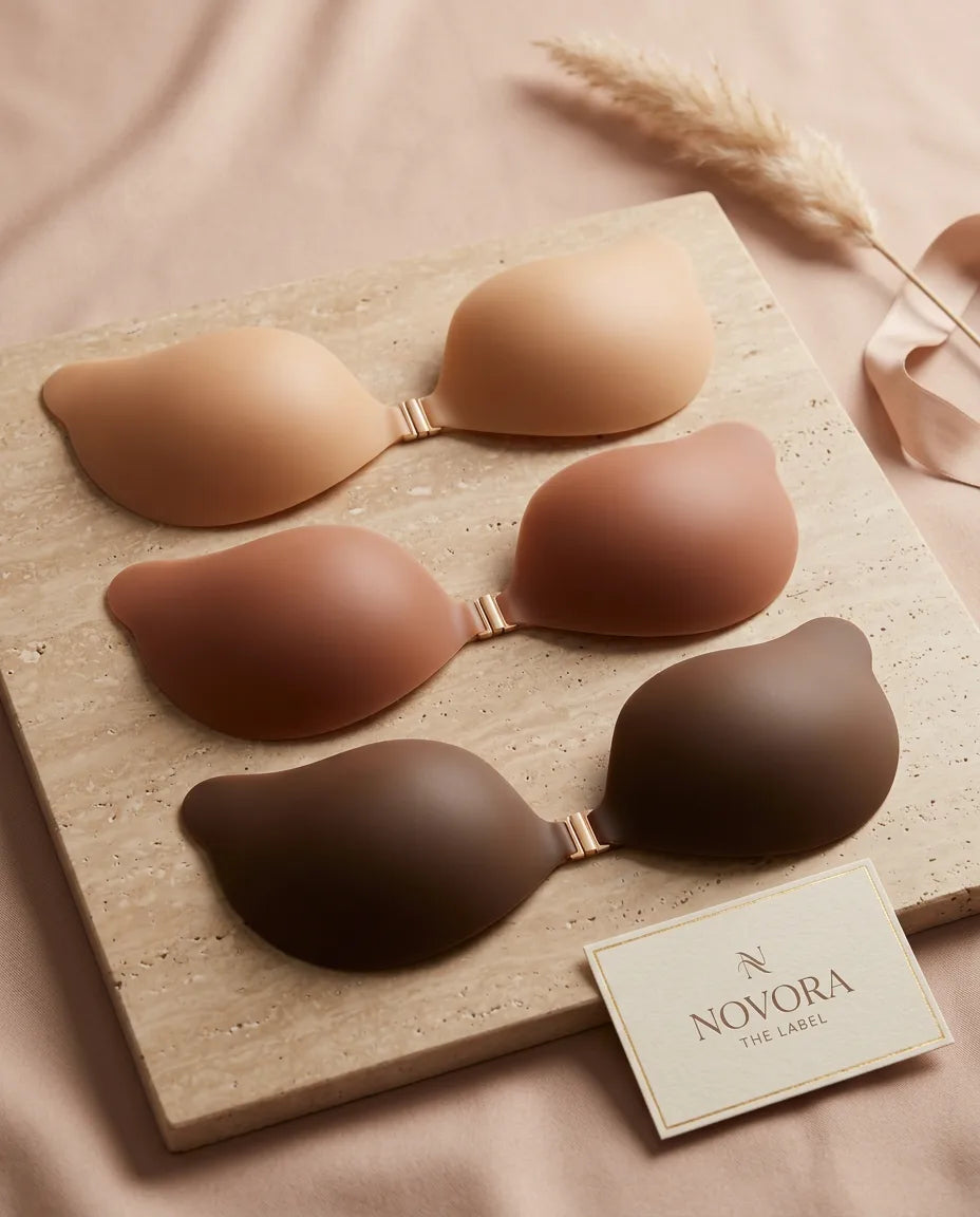 Three pairs of silicone adhesive bra cups in light nude, tan and deep brown laid on a stone board beside a Novora The Label card
