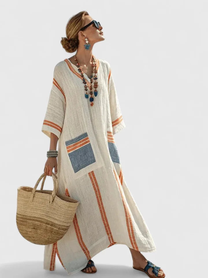 Cream maxi dress with orange stripes and denim-blue striped pocket detail, worn walking with a woven straw tote