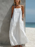White vertical-striped maxi dress with thin adjustable straps, worn against a textured grey wall by the beach