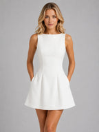 Front view of a sleeveless white A-line mini dress with a boat neckline and seamed bodice, styled with the hands in the side pockets