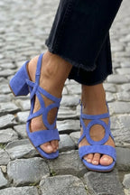 Blue suede strappy sandal with oval cut-out detail and low block heel, worn outdoors on cobblestones