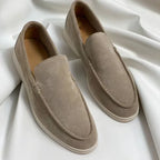 Pair of beige suede slip-on loafers with white soles shown on a draped white fabric backdrop