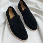 Black suede slip-on loafers with a subtle sheen and off-white soles on a white fabric background