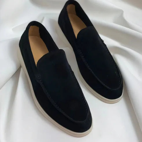 Black suede slip-on loafers with a subtle sheen and off-white soles on a white fabric background