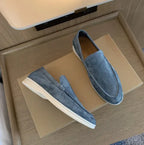 Grey suede slip-on loafers with white soles resting on a wooden shelf, one shoe on its side