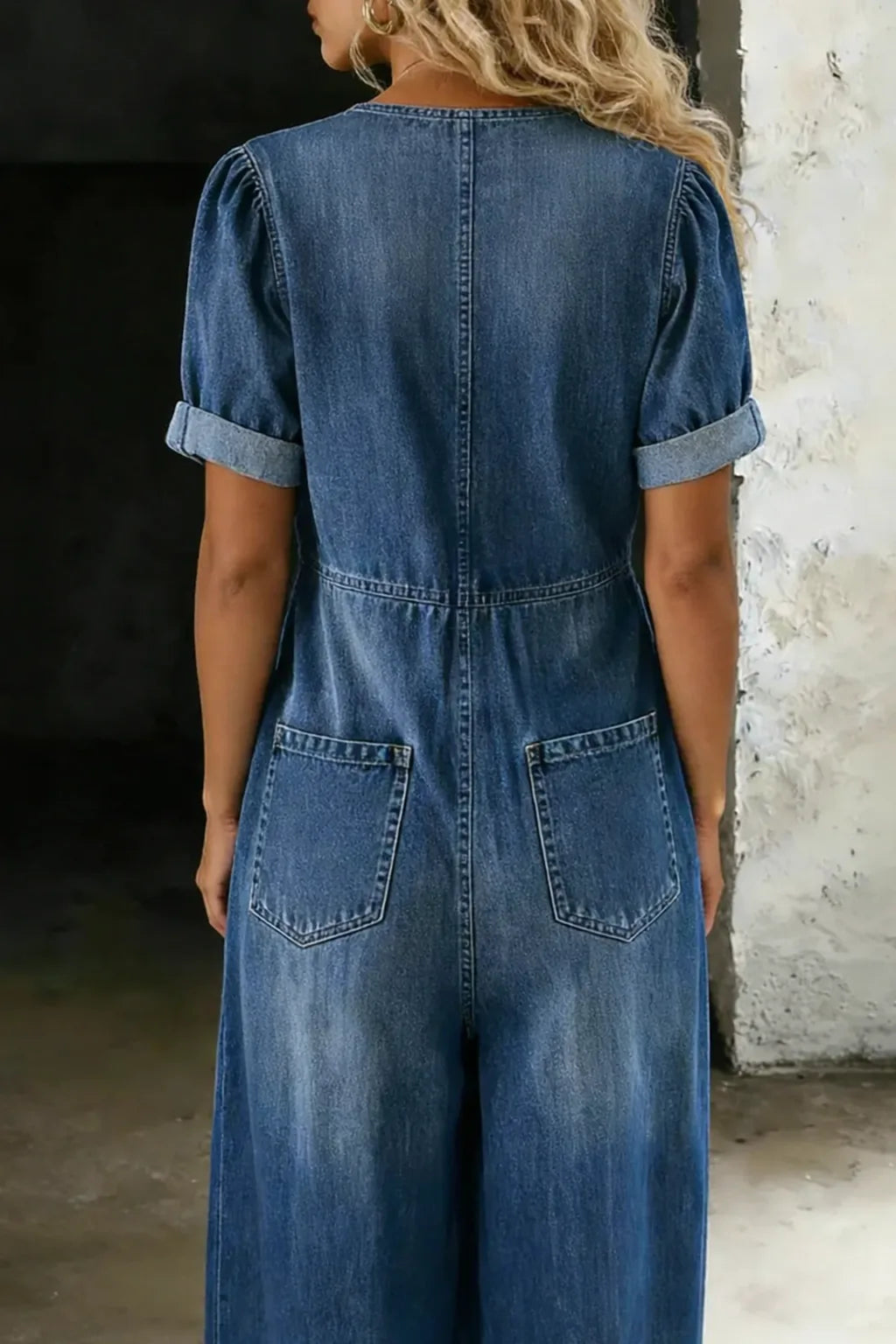 Back view of the blue denim jumpsuit showing puff sleeves, fitted waist seam and twin patch pockets