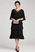 Front view of a woman in a black tiered ruffle midi dress with cape sleeves, holding a sparkly silver clutch and statement necklace