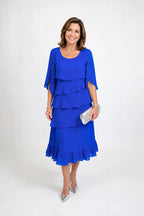 Front view of a woman in a royal blue tiered ruffle midi dress with cape sleeves, styled with a silver clutch and heels