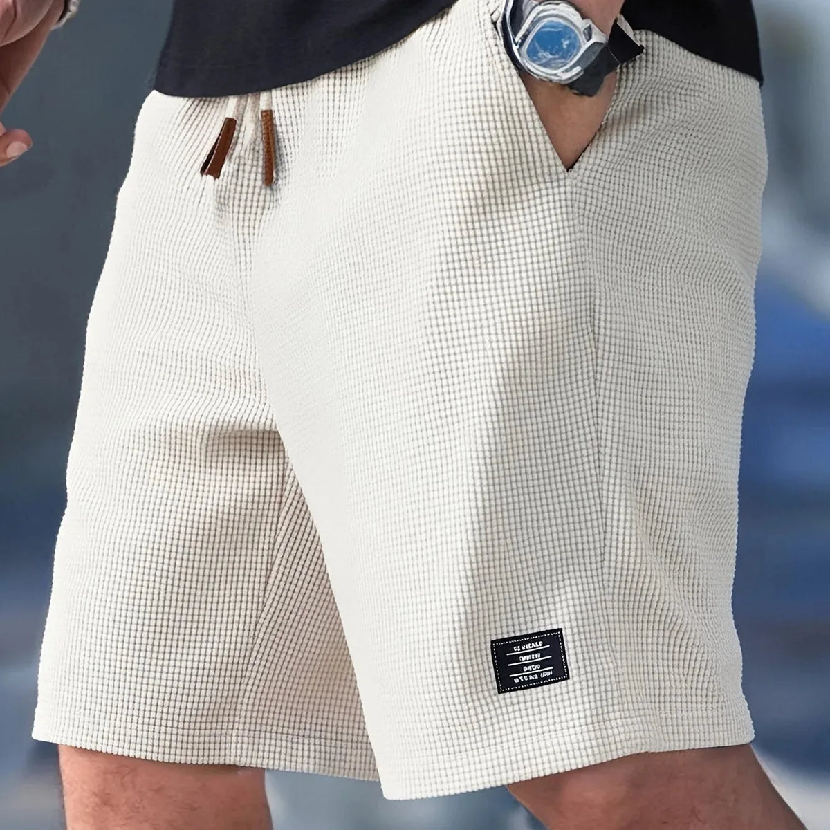 Off-white textured shorts worn with a watch and black tee, showing side pocket opening and hem patch