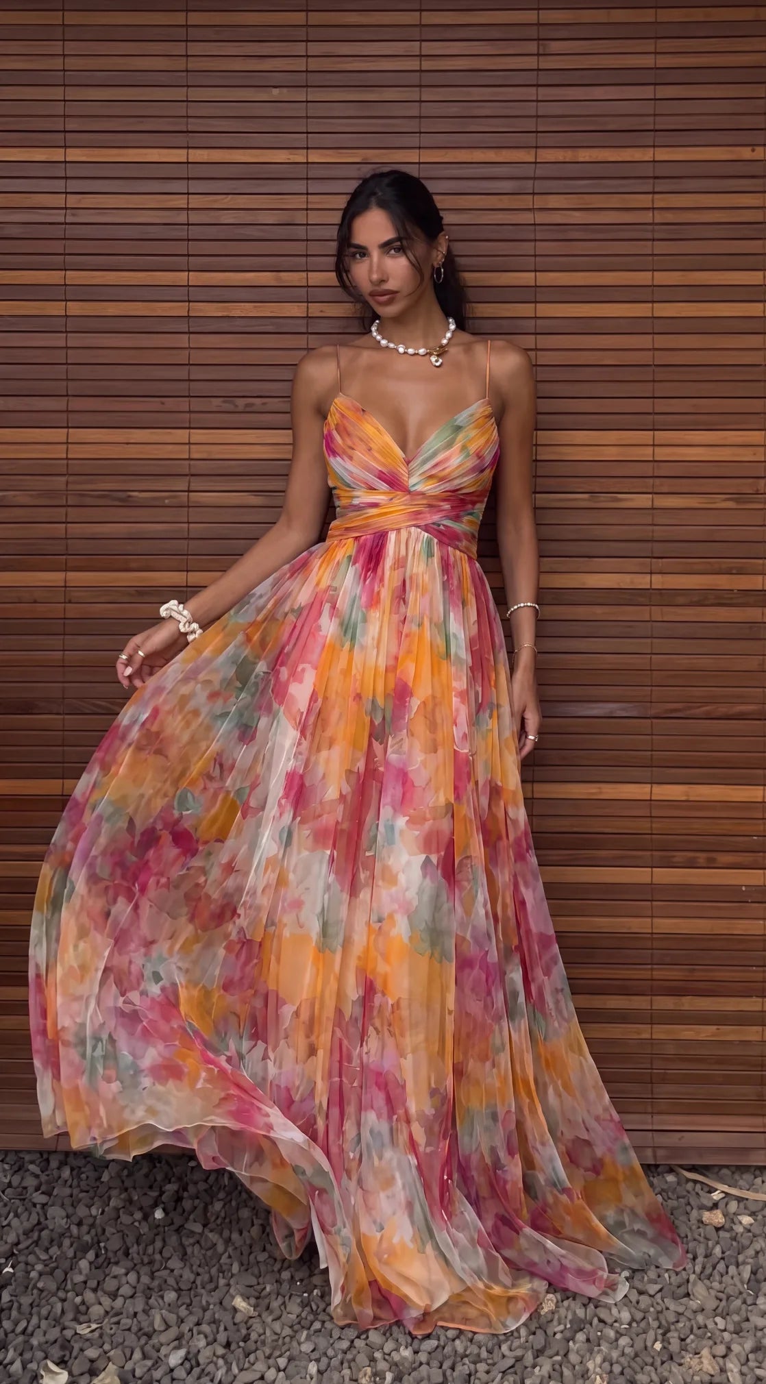 Model in a vivid orange, pink and green watercolour floral maxi dress with a V neckline, standing against a wooden slat wall