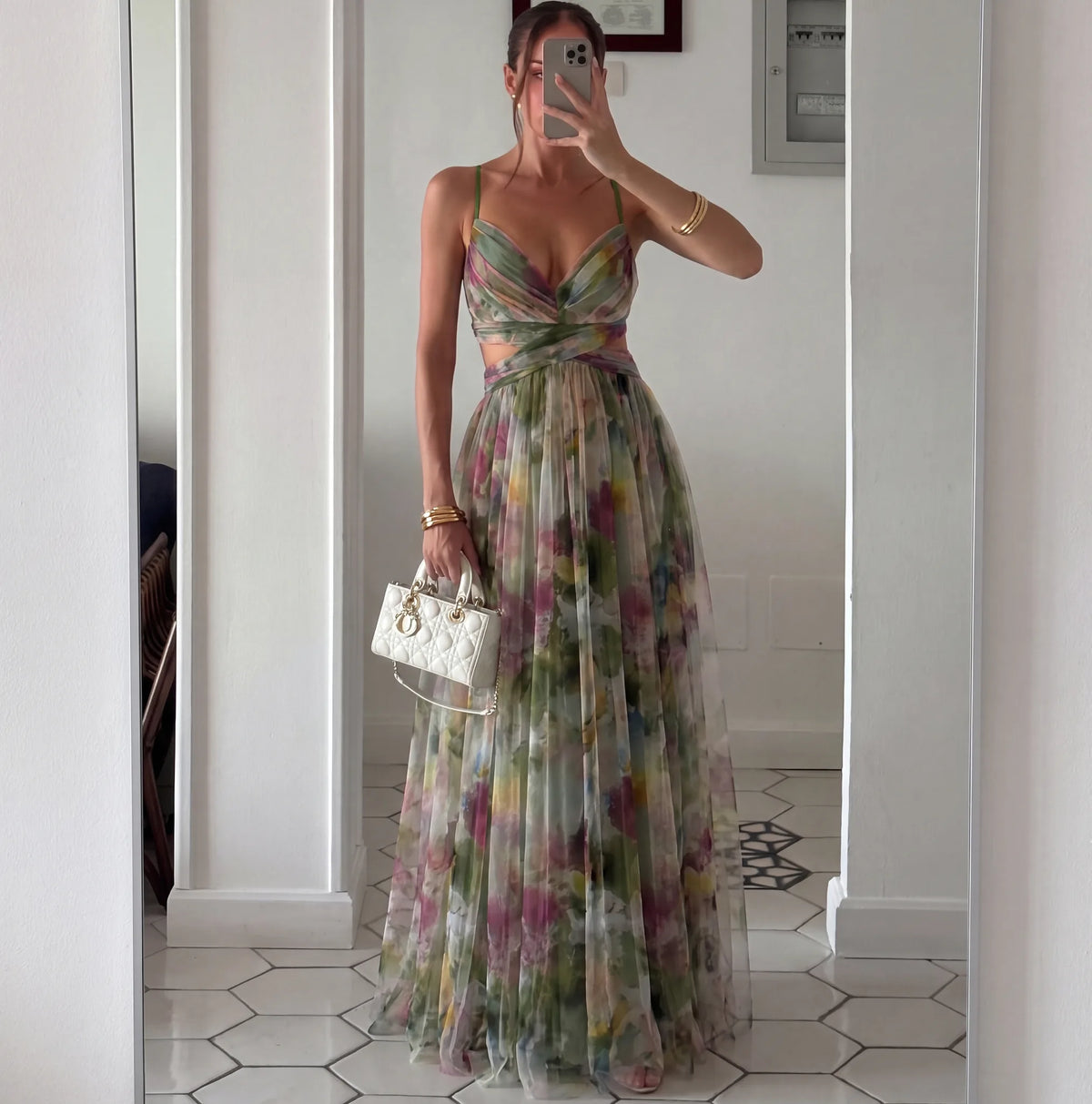 Mirror selfie of a model in the green floral maxi dress with cutout waist, pleated skirt and a white quilted handbag