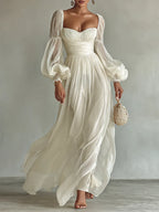 Model wearing a white chiffon maxi dress with sweetheart neckline, ruched waist and sheer balloon sleeves, holding a woven bag
