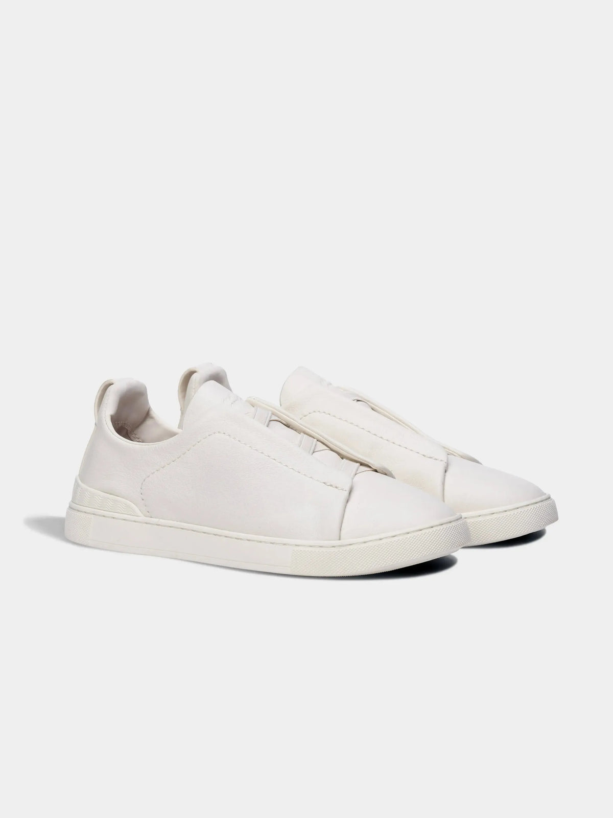 Front three-quarter view of a pair of white leather sneakers with minimalist laceless design on a light background.