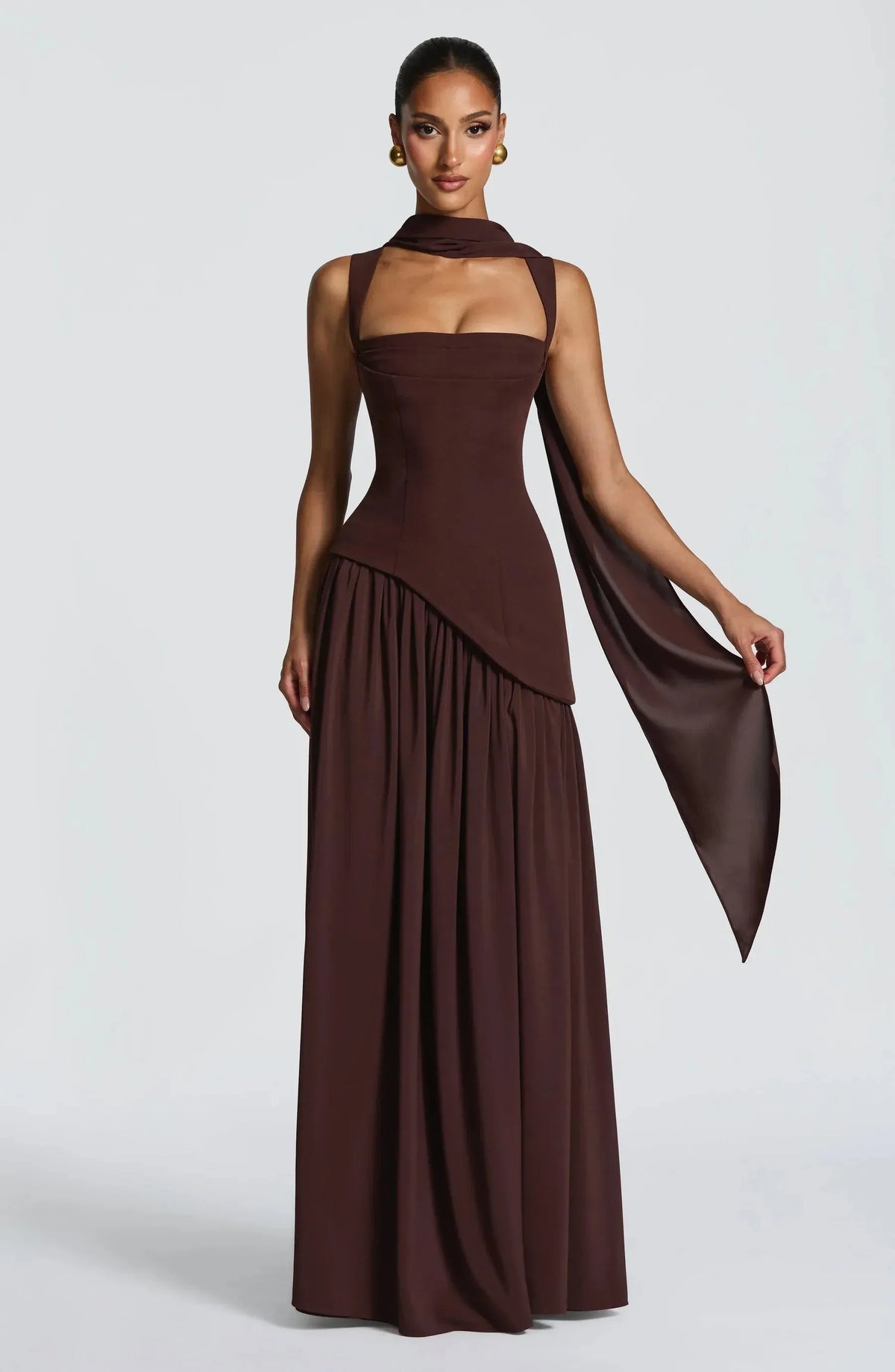Front view of model in a deep brown sleeveless maxi dress featuring a draped shoulder scarf detail and flowing skirt