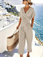 Woman wearing a beige wrap-front jumpsuit with short sleeves, cropped wide-leg pants and rolled hems, carrying a woven tote by the sea