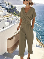 Woman wearing an olive green wrap-front jumpsuit with V-neckline, short sleeves and cropped wide-leg pants on a sunlit cliffside path