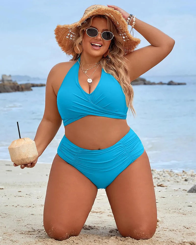 Plus-size model kneeling on sand in a bright blue wrap halter bikini with ruched high-waisted bottom