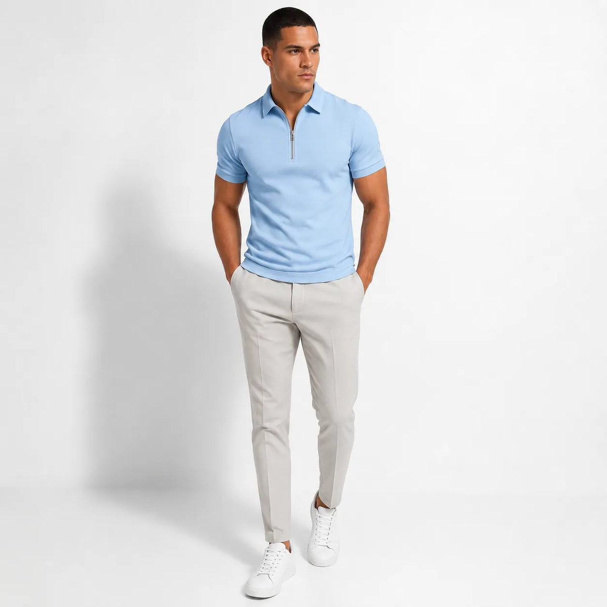 Full-body shot of a man wearing a light blue zip-neck polo shirt tucked into grey tailored trousers with white sneakers